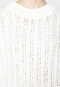 Weißer Strickpullover mit einer gerippten Struktur, der vertikale Streifen und einen weiten Ausschnitt aufweist. Der Stoff wirkt leicht und atmungsaktiv.