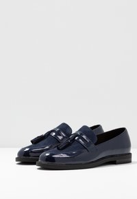 Mørkeblå loafers laget av glansfull patentlær og semsket skinn, med en dekorativ dusk og en svart såle med lav hæl.