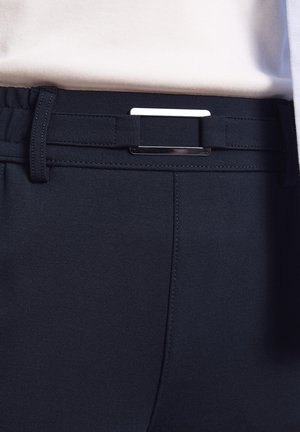 Nahaufnahme von schwarzen Hosen mit einem elastischen Gürtel mit rechteckiger silberner Schnalle, kombiniert mit einem weißen Oberteil.