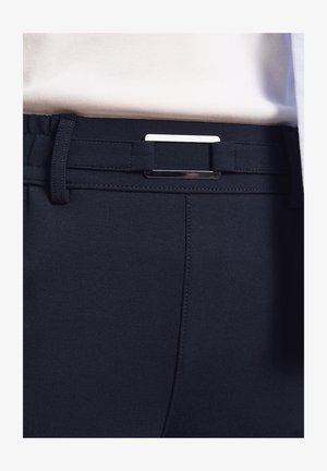 Nahaufnahme von schwarzen Hosen mit einem elastischen Gürtel mit rechteckiger silberner Schnalle, kombiniert mit einem weißen Oberteil.