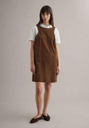 Frau steht und trägt ein braunes ärmelloses Kleid über einem weißen kurzärmligen Hemd mit Kragen und dunkelbraunen Ballerinas vor einem einfarbigen Hintergrund.