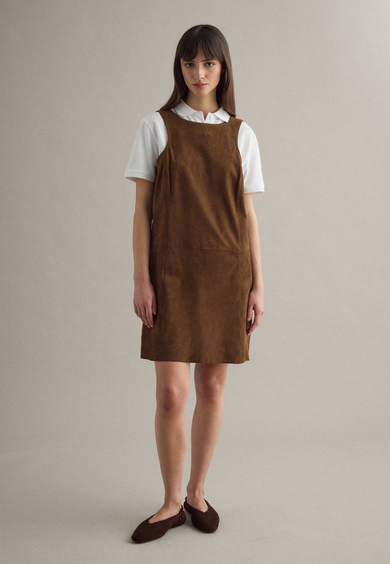 Frau steht und trägt ein braunes ärmelloses Kleid über einem weißen kurzärmligen Hemd mit Kragen und dunkelbraunen Ballerinas vor einem einfarbigen Hintergrund.