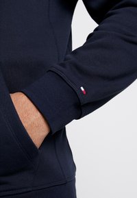 Navy blue sweatshirt with a smooth texture, featuring a small embroidered red, white, and blue logo on the cuff and a front pocket.
