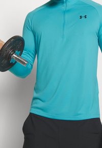 Helleblaue langärmlige Sportshirt mit einem halbhohen Reißverschlusskragen, mit einem schwarzen Logo auf der Brust. Hält eine schwarze Gewichtsplatte.