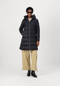 Black puffer coat with a hood, featuring a zipper front and vertical stitching. Paired with beige cargo pants and black ankle boots.