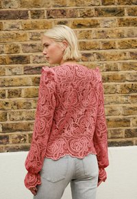 Roza čipkasta bluza z dolgimi rokavi, ki vsebuje zapletene cvetlične vzorce in nagubane poudarke, v kombinaciji s svetlo sivimi oprijetimi kavbojkami.