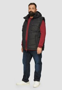 Schwarze gepolsterte Weste mit Kapuze, ausgestattet mit Fronttaschen und einem Reißverschluss. Tragen über einem roten Langarmshirt und kombiniert mit dunkler Jeans und weißen Sneakers.