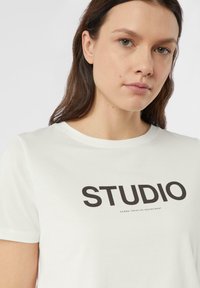 Biały bawełniany T-shirt z krótkim rękawem, z dużym czarnym napisem "STUDIO" na przodzie oraz mniejszym tekstem poniżej. Gładka faktura.