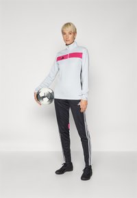 Light gray long-sleeve top with a pink panel and black accents, paired with black joggers featuring pink details and white stripes; holding a soccer ball.
