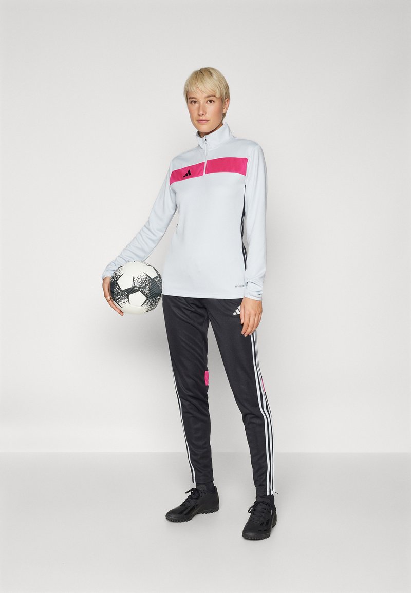Lichtgrijs langemouwtop met een roze paneel en zwarte accenten, gecombineerd met zwarte joggingbroeken met roze details en witte strepen; houdt een voetbal vast.