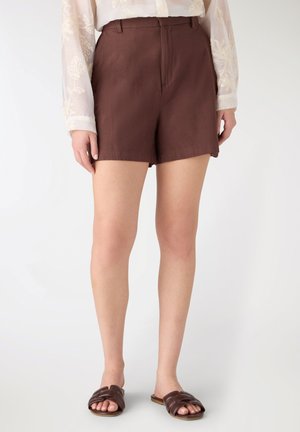 Donna che indossa pantaloncini marroni a vita alta con passanti per cintura, abbinati a sandali bassi marrone scuro, in piedi su sfondo bianco.