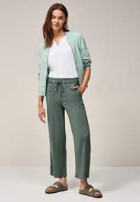 Veste bomber vert clair sur un haut blanc, associée à un pantalon large vert foncé et des sandales marron avec une boucle. Texture du tissu lisse.