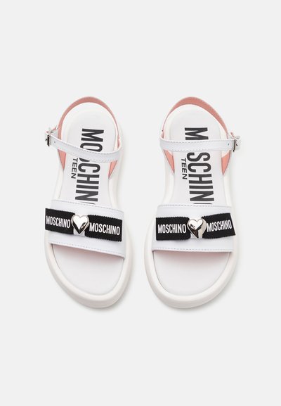 Sandalias blancas con una plantilla rosa, con correas elásticas negras que llevan el texto "MOSCHINO" y un adorno en forma de corazón plateado. Correa de tobillo ajustable.
