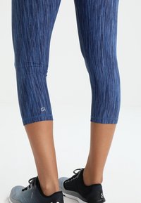 Blue, capri-length leggings featuring a vertical striped pattern and a small logo near the hem. Paired with black athletic shoes.