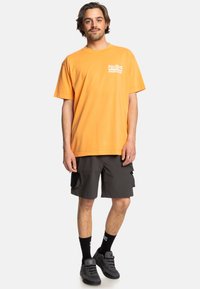 Orange cotton t-shirt with front logo, paired with dark gray cargo shorts, black crew socks, and black shoes. Model stands front-facing.