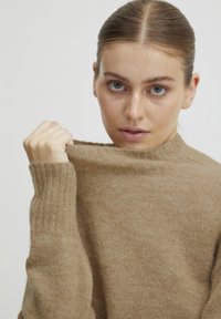 Pull marron clair avec un col montant et des poignets côtelés. Le tissu semble doux avec une texture lisse. Le mannequin tient délicatement le col.