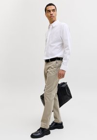 Mann som bærer hvit skjorte, beige bukser og svarte sko, holder en svart duffelbag og står mot en ensfarget lys bakgrunn.