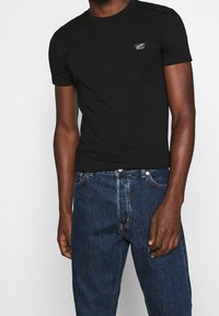 Schwarzes T-Shirt aus Baumwolle, mit Rundhalsausschnitt und kurzen Ärmeln. Ein kleines Logo-Patch befindet sich auf der linken Brustseite.