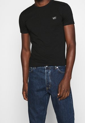 Schwarzes T-Shirt aus Baumwolle, mit Rundhalsausschnitt und kurzen Ärmeln. Ein kleines Logo-Patch befindet sich auf der linken Brustseite.