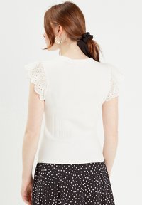 Haut en tricot côtelé blanc avec des manches courtes en dentelle. Le dos présente un col rond, associé à une jupe à pois noir et blanc.