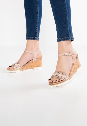 Beige wedgesandal med korkhæl, der har tre dekorative remme med metalliske detaljer og en justerbar ankelrem.