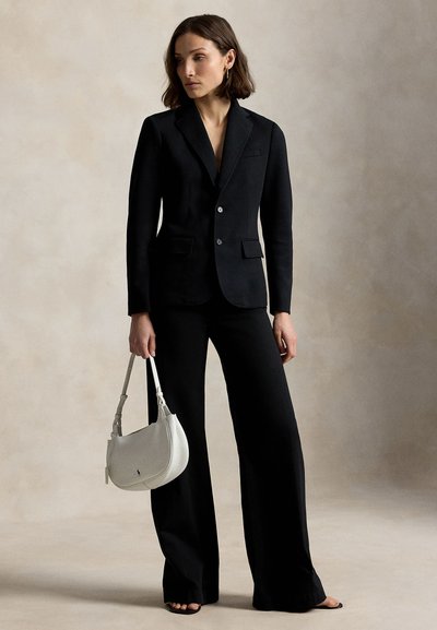 Vrouw draagt een zwart blazer en broek met wijde pijpen, houdt een witte handtas vast, staand tegen een effen lichte achtergrond.