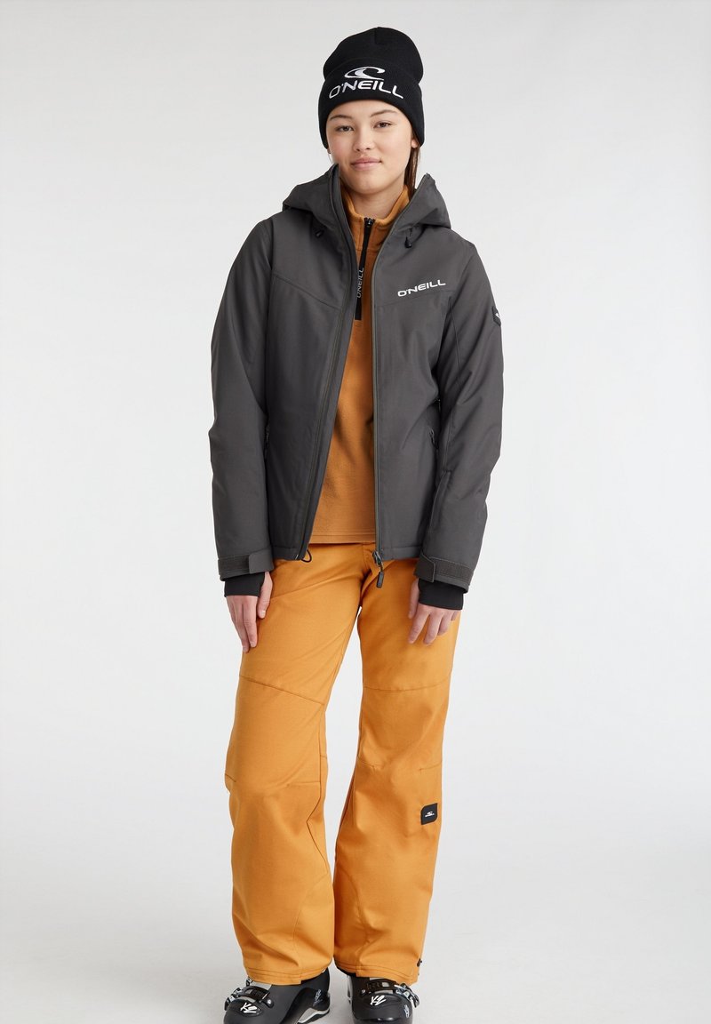 Szara kurtka z zamkiem błyskawicznym i kapturem, pomarańczowe spodnie oraz czarna czapka. Stylizowana na odzież outdoorową; cechy to oddychający materiał i miękka tekstura.