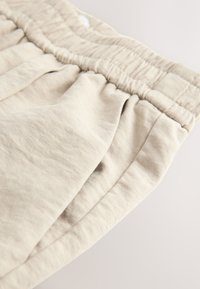 Heledast beežid puuvillased püksid elastse vöökohaga, millel on pehme tekstuur ja vöökohal voltimine, vaadatuna nurga alt.