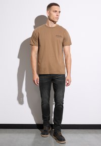 Brun bomull t-shirt med korta ärmar har en liten svart "CUSTOM MADE" logotyp på bröstet. Kombineras med mörkgrå slim-fit jeans och stövlar.