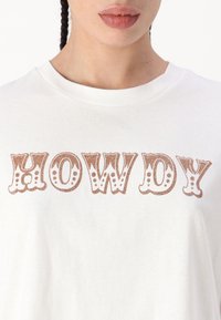 Bílý bavlněný tričko s velkým, třpytivým hnědým nápisem "HOWDY" napsaným hravým písmem. Kulatý výstřih, krátké rukávy.