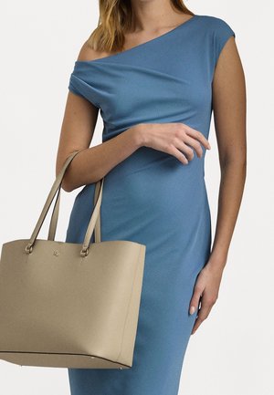 Sac fourre-tout beige au design structuré, texture en cuir lisse, avec deux poignées, tenu contre une robe bleue à encolure drapée.