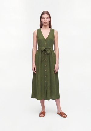 Vrouw staat recht voor zich uitkijkend, draagt een mouwloze olijfgroene midi-jurk met knopen en een gestrikte tailleband, gecombineerd met bruine platte sandalen.