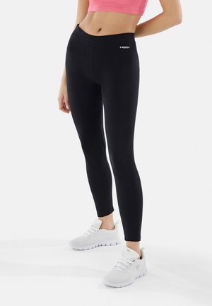 Leggings neri in tessuto elasticizzato, con vita alta e un piccolo logo bianco sul lato, abbinati a scarpe da ginnastica bianche.