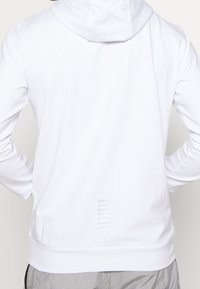 Bílá mikina s kapucí s hladkým povrchem, přiléhavým střihem a nenápadnými detaily na spodní části zad s horizontálními řeznými liniemi.