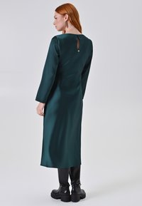 Abito lungo in satin verde scuro con maniche lunghe e dettaglio a apertura a chiave sulla schiena. Indossato con stivali neri fino al ginocchio e tocchi minimali.