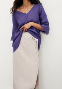 Haut en lin violet avec un profond décolleté en V et des manches à franges, associé à une jupe portefeuille beige clair avec une fente latérale et un détail noué.