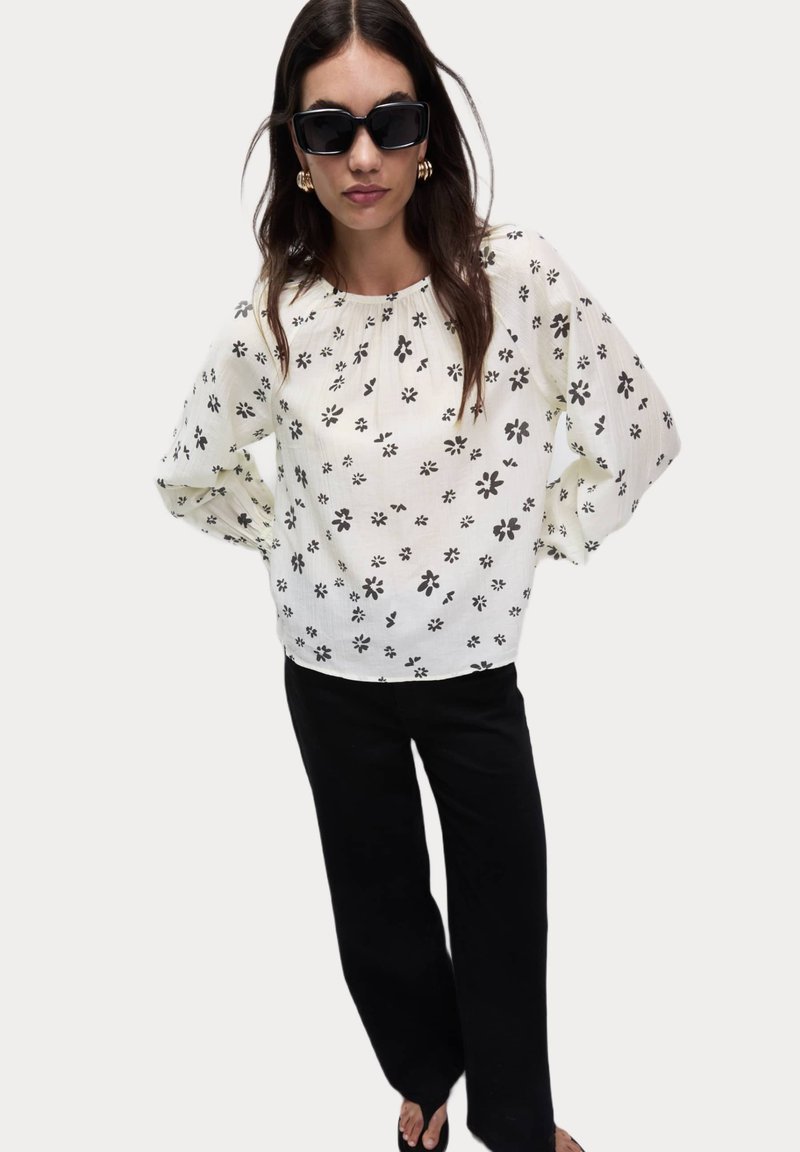 Blusa bianca con motivo floreale nero, maniche lunghe e a sbuffo, scollo arricciato; abbinata a pantaloni neri larghi e occhiali da sole scuri.