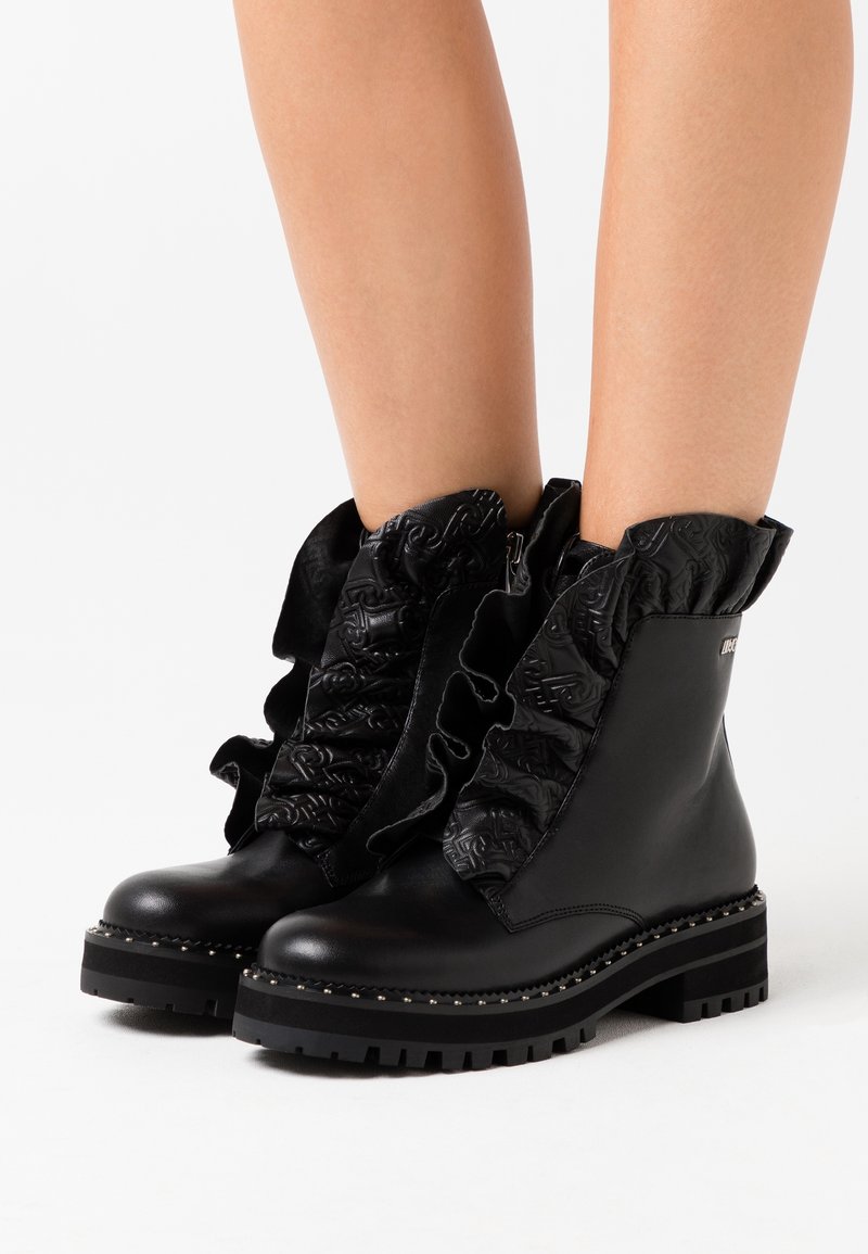 Botas de tobillo de cuero negro con detalles rizados texturizados a lo largo de los laterales, suela gruesa y acentos de tachuelas alrededor de la base.