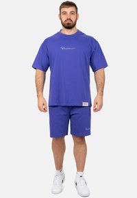Ensemble t-shirt et short en coton violet à coupe ample. Comprend un logo sur la poitrine et l'ourlet ; associé à des baskets blanches.
