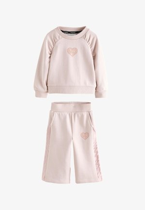 Småbarn iført lyserød sweatshirt og vide bukser med hjerte-designs, smilende og kigger let opad mod en ensfarvet baggrund.