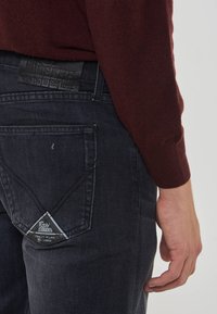 Uomo che indossa jeans di denim blu scuro con un'etichetta di pelle sulla vita posteriore e un maglione a maniche lunghe bordeaux.