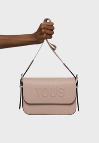 Borsa a tracolla in pelle rosa con chiusura a pattina, logo "TOUS" impresso, tracolla regolabile e dettagli in metallo argentato. Texture liscia, forma rettangolare.