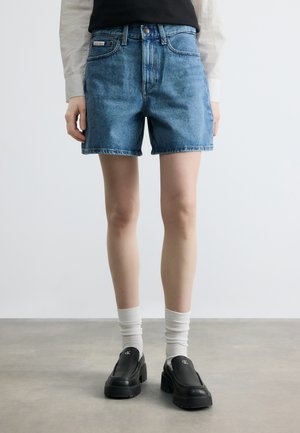 Isik, kes kannab keskmise sinise teksapükse, valgeid sokke ja musti libisevaid kingi valge logoga, halli põranda taustal.