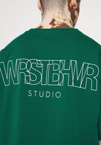 Grön bomullströja med stora vita bokstäver på baksidan som läser "WRSTBHV Studio." Rund halsringning, avslappnad passform, minimala texturdetaljer.