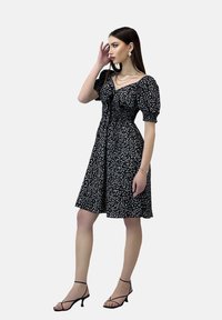 Vestido negro con patrón floral blanco, mangas cortas abullonadas, corsé ajustado y falda acampanada. Combinado con zapatos de tacón negros con tiras y complementos.