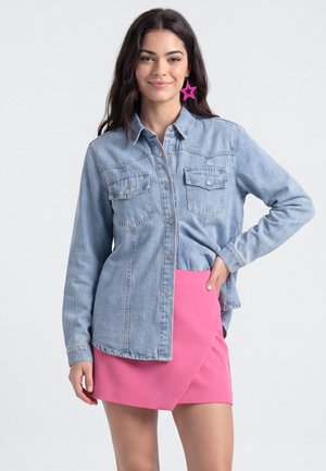 Junge Frau mit langen dunklen Haaren trägt eine hellblaue Jeansjacke, einen knallpinken Rock und pinke sternförmige Ohrringe, posiert mit der Hand in der Hüfte.