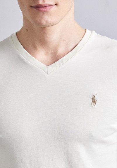 T-shirt à col en V et manches courtes en coton beige clair. Présente un petit logo brodé sur la poitrine gauche. Texture lisse, design minimal.