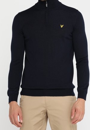 Uomo che indossa un maglione nero aderente con cerniera a un quarto e un piccolo logo di uccello giallo sul petto, abbinato a pantaloni beige.