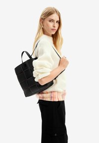 Černá taška přes rameno s texturovaným povrchem a mřížkovým vzorem, s kontrastním oranžovým prošíváním. Ucho je připevněno a nosí se na rameni.