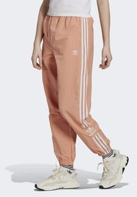 Béžové nylonové kalhoty s bílými pruhy po stranách, elastickým pasem a zúženými kotníky. Na přední straně je logo Adidas.